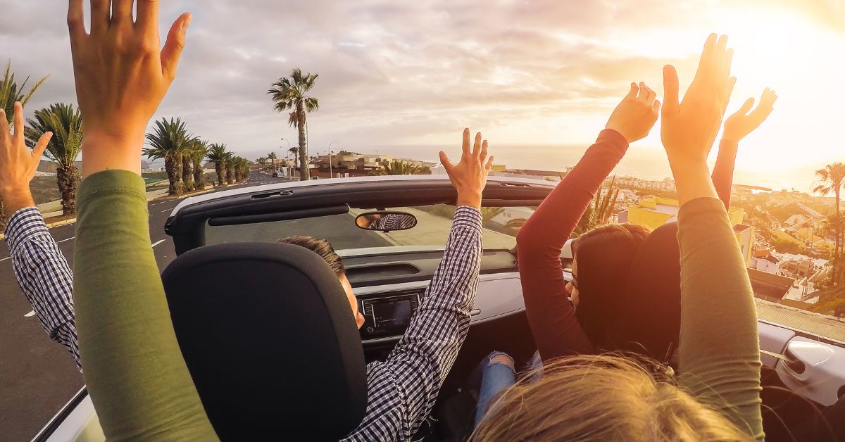 People with their arms up, driving in a car with the sunset in the distance