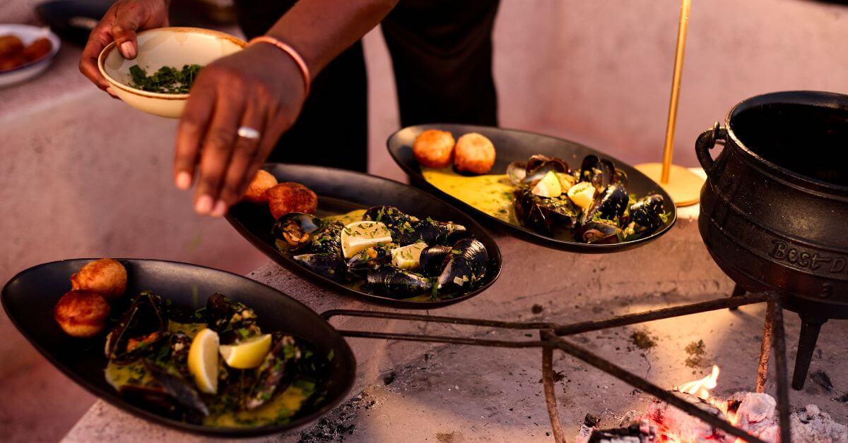 a chef dressing mussels served on black plates outside with a fire and a potjie pot next to it.